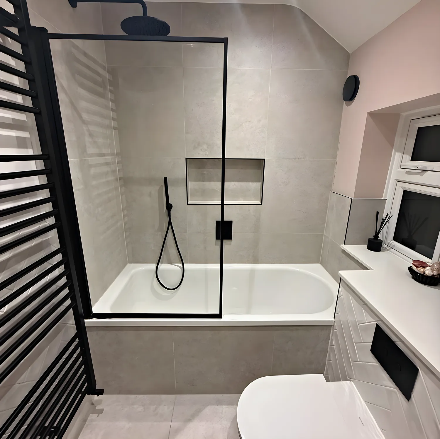 A full size bath with a black framed screen, grey tiles, a tiled niche and a black ladder towel rail