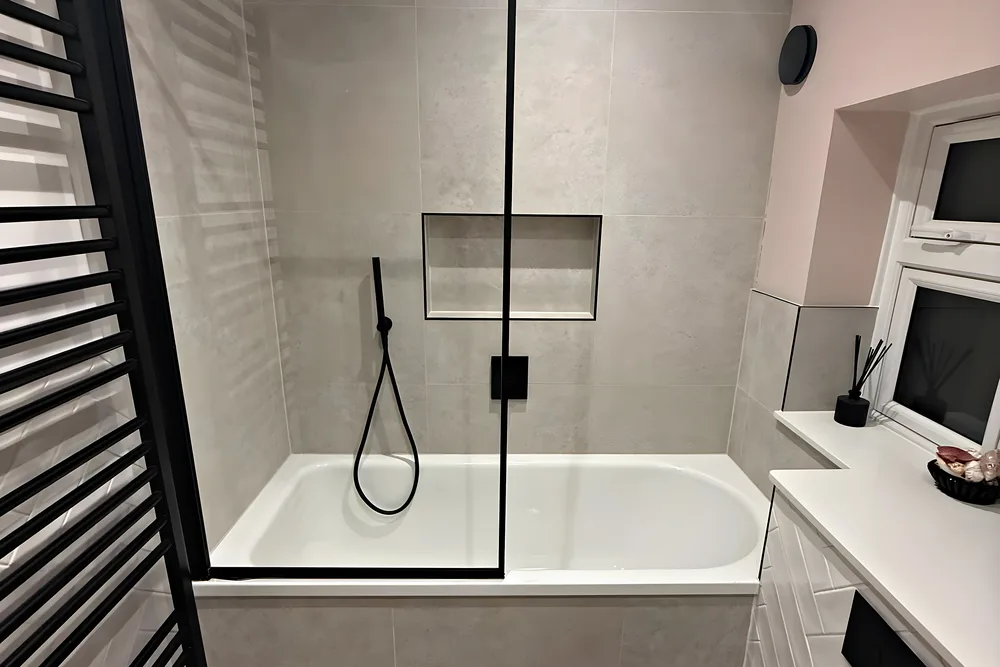 A full size bath with a black framed screen, grey tiles, a tiled niche and a black ladder towel rail