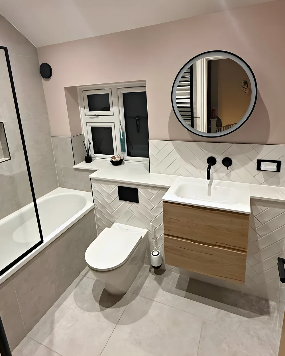 A bathroom with baby pink walls, a round black mirror, an oak vanity unit and a wall hung toilet
